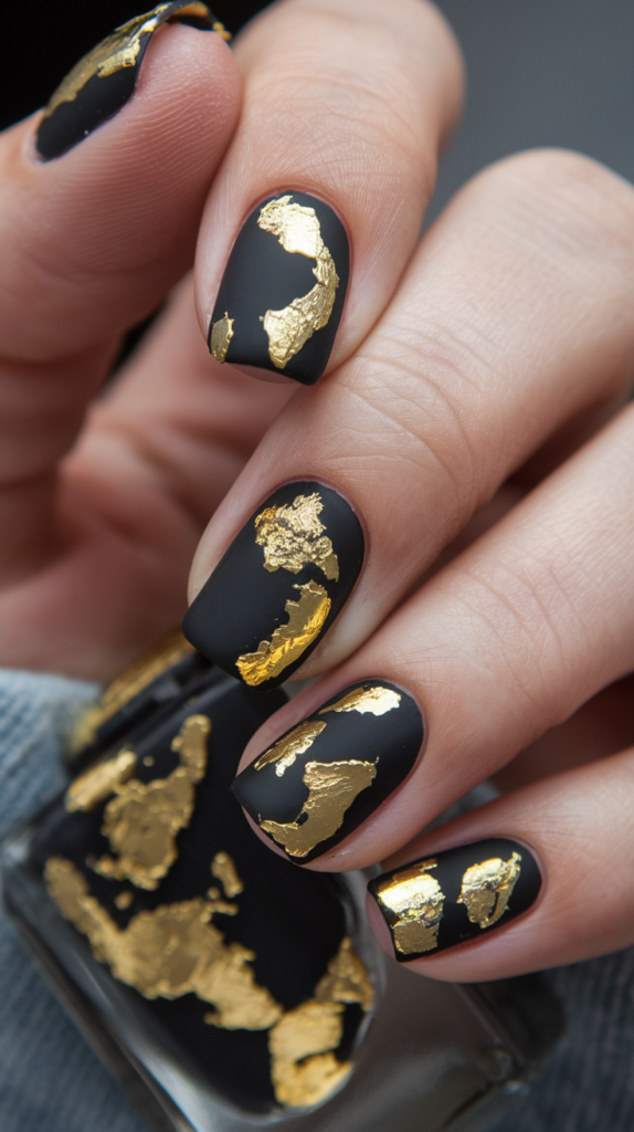 Matte black nails adorned with scattered pieces of shiny gold leaf.
