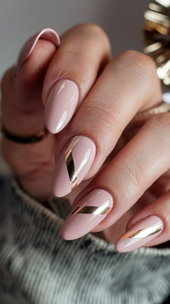 Close-up of almond-shaped nails painted in soft blush pink with sleek diagonal gold foil stripes, finished with a glossy top coat.